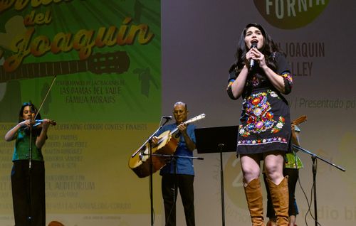 A woman in a black dress, embroidered with colorful flowers, singing on a stage. Three musicians play instruments behind her.