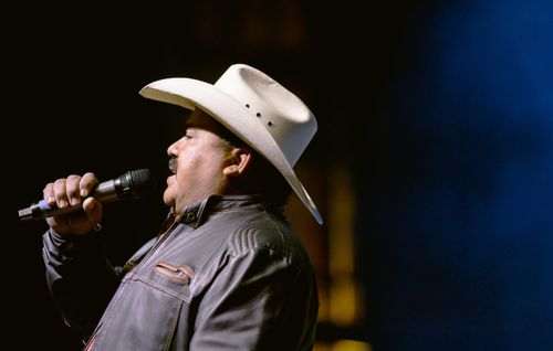 A close-up photo, taken from the side, of Rodolfo Carranza holding a mic and singing. He wears a tan cowboy hat and a dark brown leather jacket.