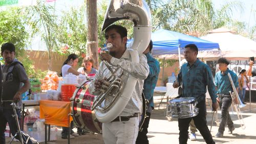 A man dressed all in white and silver plays a white and silver sousaphone tuba. In the background, members of a band carry their drums and brass instruments, while women set up refreshments.