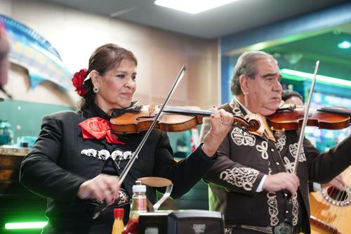 Inside a restaurant, two mariachis, a woman and a man in black suits, play violin.