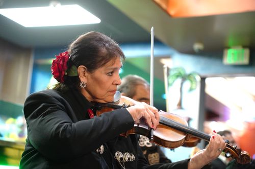 A close-up photo of a woman mariachi, Carmen Dias, leaning forward slightly as she plays violin inside a brightly lit restaurant.