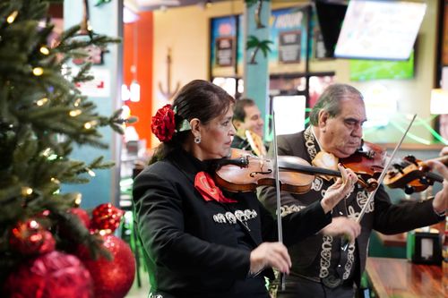 Inside a restaurant, two mariachis, a woman and a man in black suits, play violin next to a Christmas tree.