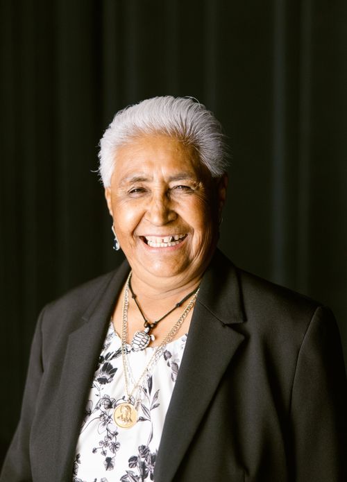 A portrait of a woman with short, white hair and a black blazer smiling widely in front of a black curtain.