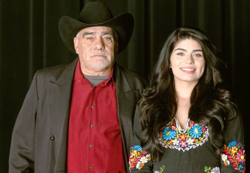 A portrait of a serious-faced man and a smiling young woman, who is his daughter, standing in front of a black curtain.