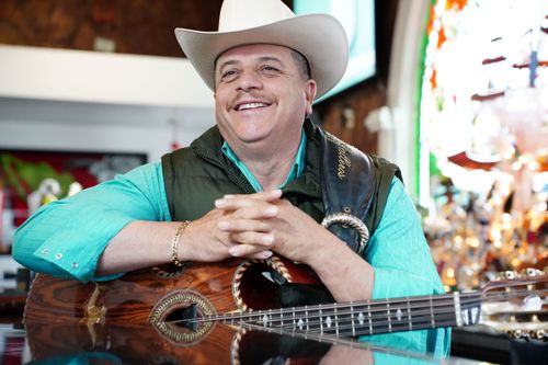 A man in a white cowboy hat, a green puffer vest, and a bright teal shirt sits at a restaurant bar and smiles, while resting his arms on a guitar.