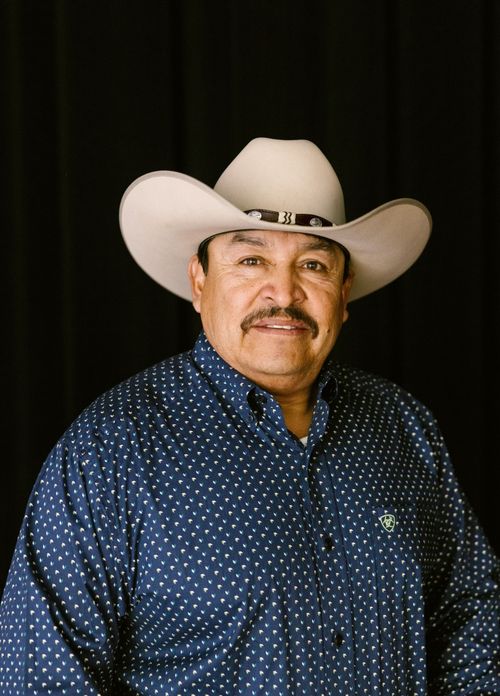 A portrait of a mustached man standing in front of a black curtain. He wears a blue shirt with white polka dots, along with a beige cowboy hat.