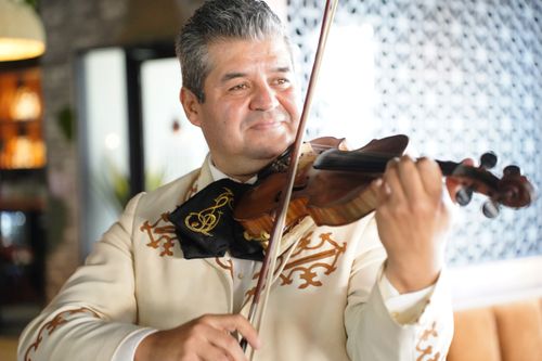 A close-up photo of a gray-haired man in an off-white mariachi outfit and black tie smiling while playing violin.