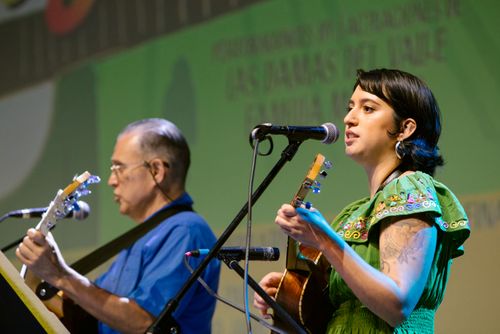 A man in a blue shirt and a woman in a green shirt sing together on stage. The man plays guitar; the woman, vihuela.