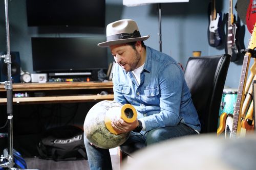Carlos Rodríguez, a man sitting in a small room filled with instruments, holds an Udu drum from the Igbo tribe in Nigeria, West Africa. It is hollow and gourd-shaped.
