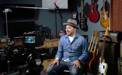 A camera on a tripod points toward Carlos Rodríguez, a man sitting in a small room filled with instruments.