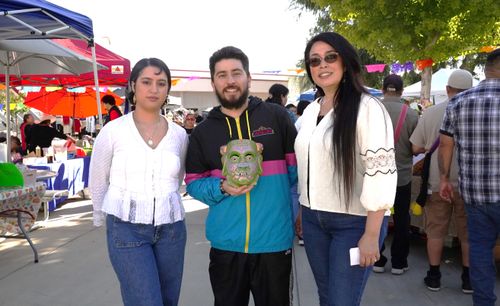 At an outdoor festival, Gabriel Gutierrez, a young man holding a decorative mask, poses for a photo. To his right stands a short-haired woman, and to his left smiles another woman with long hair.