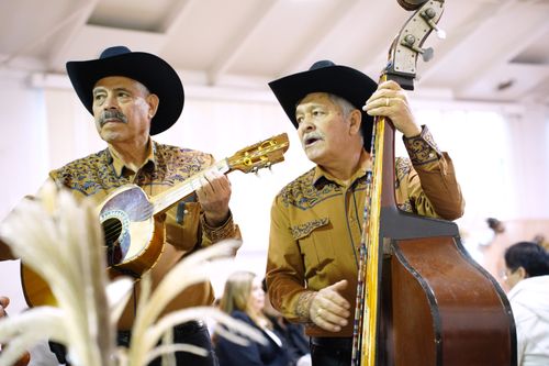 Two men in matching yellow button-down shirts and black cowboy hats play instruments inside a white room. On the left is a guitarist. On the right is Jesús Silva with an upright bass.