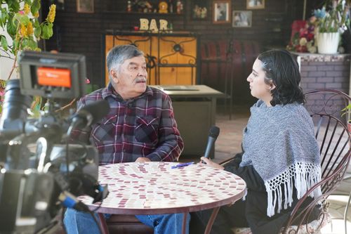 A camera points toward a grey-haired man and a short, dark-haired woman sitting at a round patio table. The man is Jesús Silva, being interviewed by Xochitl Morales outdoors.
