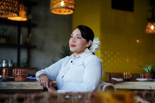 Marilyn Rodriguez, a woman in a white shirt with a white flower in her hair, leans against a table in a restaurant.