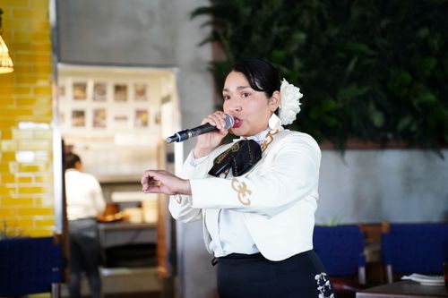 Marilyn Rodriguez, wearing a white flower in her hair, sings into a handheld microphone.