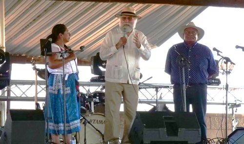 A woman, Natalia Bautista Chávez, and two men stand on a stage beneath a metal-roofed pavilion.