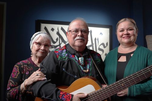 A balding man, Stanley Lucero, holds a guitar, looking toward the viewer. Holding his arm and standing to his right is smiling, white-haired Yolando Lucero; to his left is another woman: their daughter Nora Guillén.