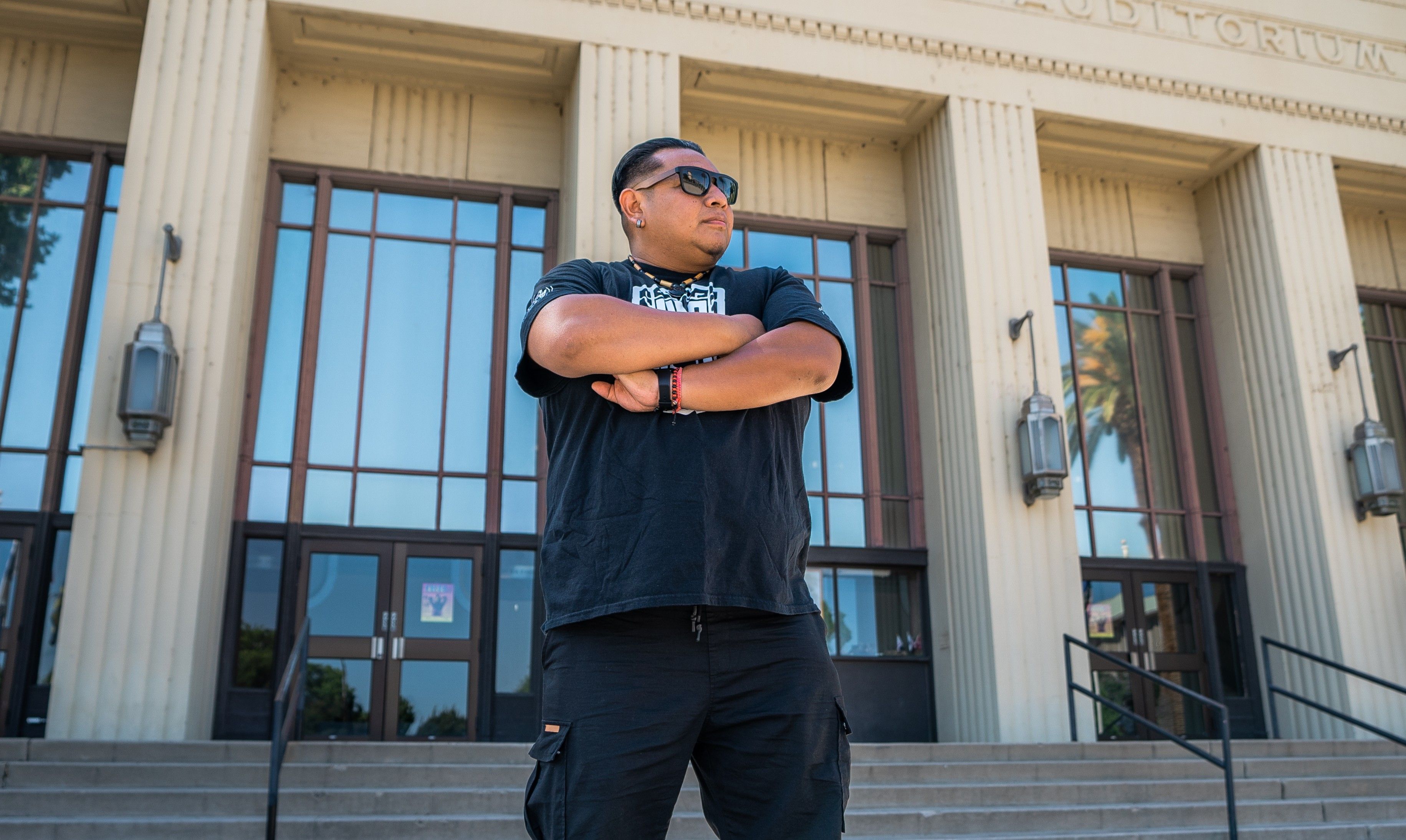 Miguel Villegas Ventura, a man in a black t-shirt and sunglasses, crosses his arms and looks off into the distance outside a large building with tall windows.