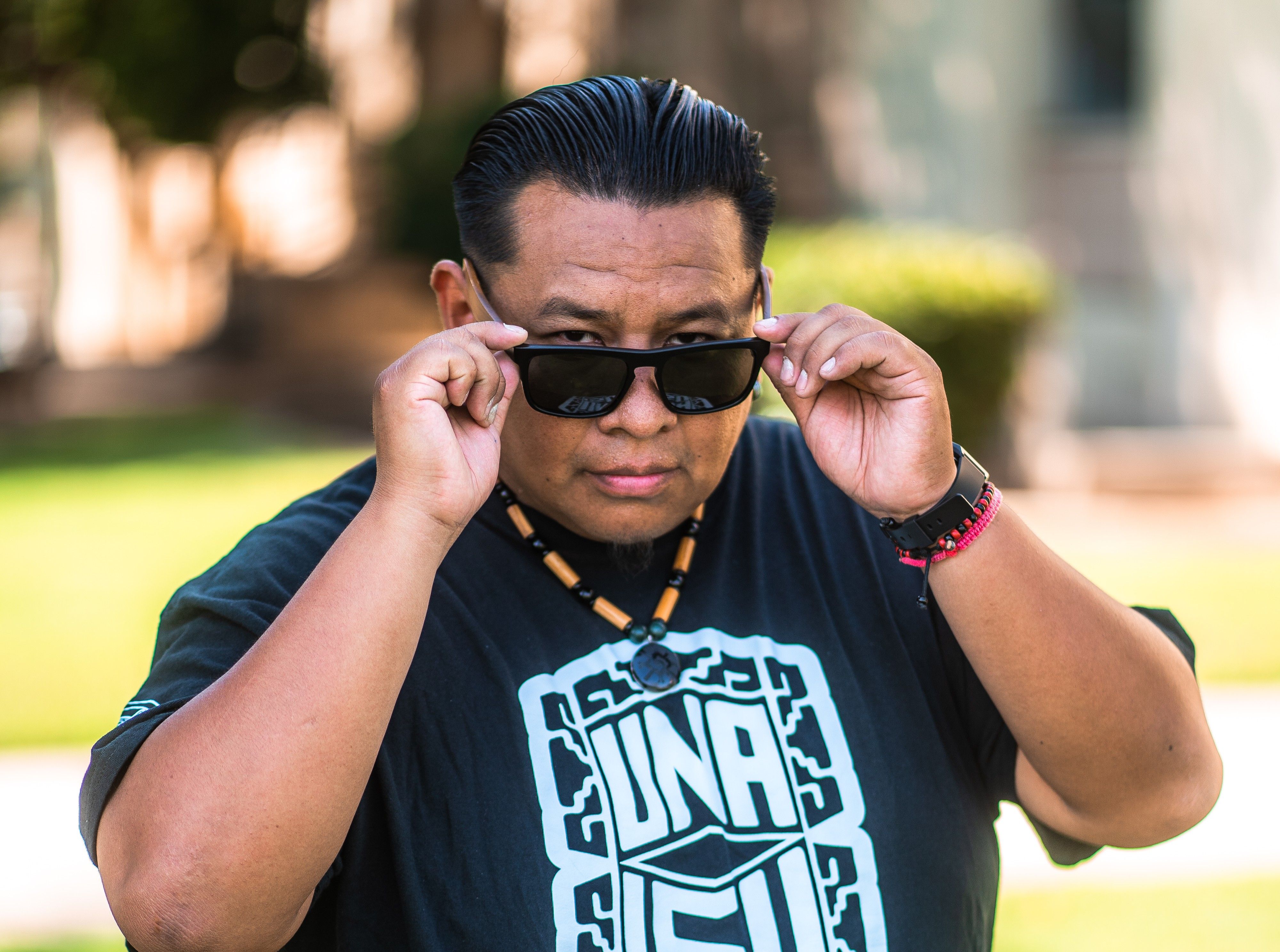A man in a black t-shirt which reads UNAISU in white, as well as a beaded necklace, lowers his sunglasses slightly to look at the camera.