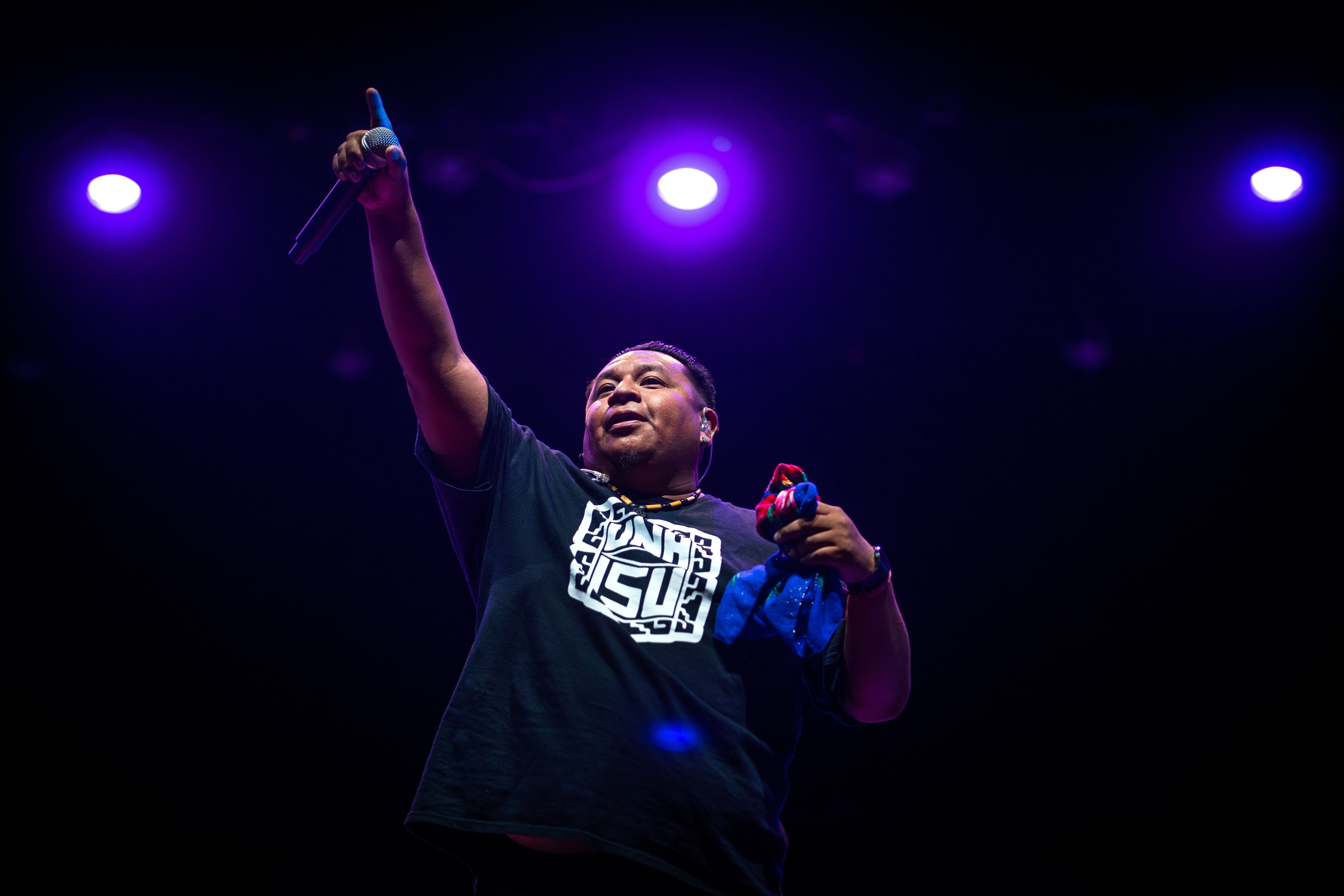 A photo taken from a low angle of a man in a black t-shirt performing beneath blue stage lights.
