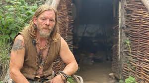 Will Lord sitting beside a primitive woven shelter for The Survival Debrief Podcast episode about Stone Age survival and flint knapping.