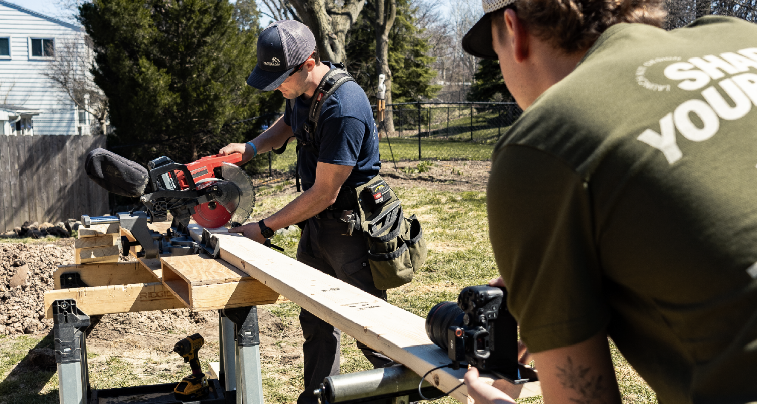 Brendan Mckellar from Mckellar Construction wearing a cap using a miter saw to cut a long wooden plank outdoors while Daniel Bremmer from Launch Kit films with a camera.