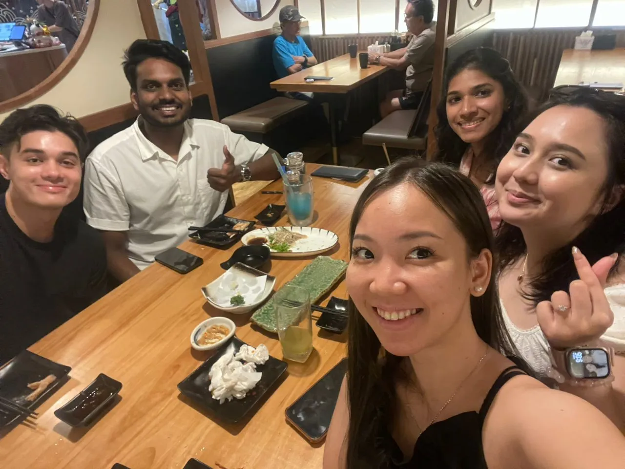 Five friends smiling and sitting around a wooden table with empty plates and drinks in a restaurant.