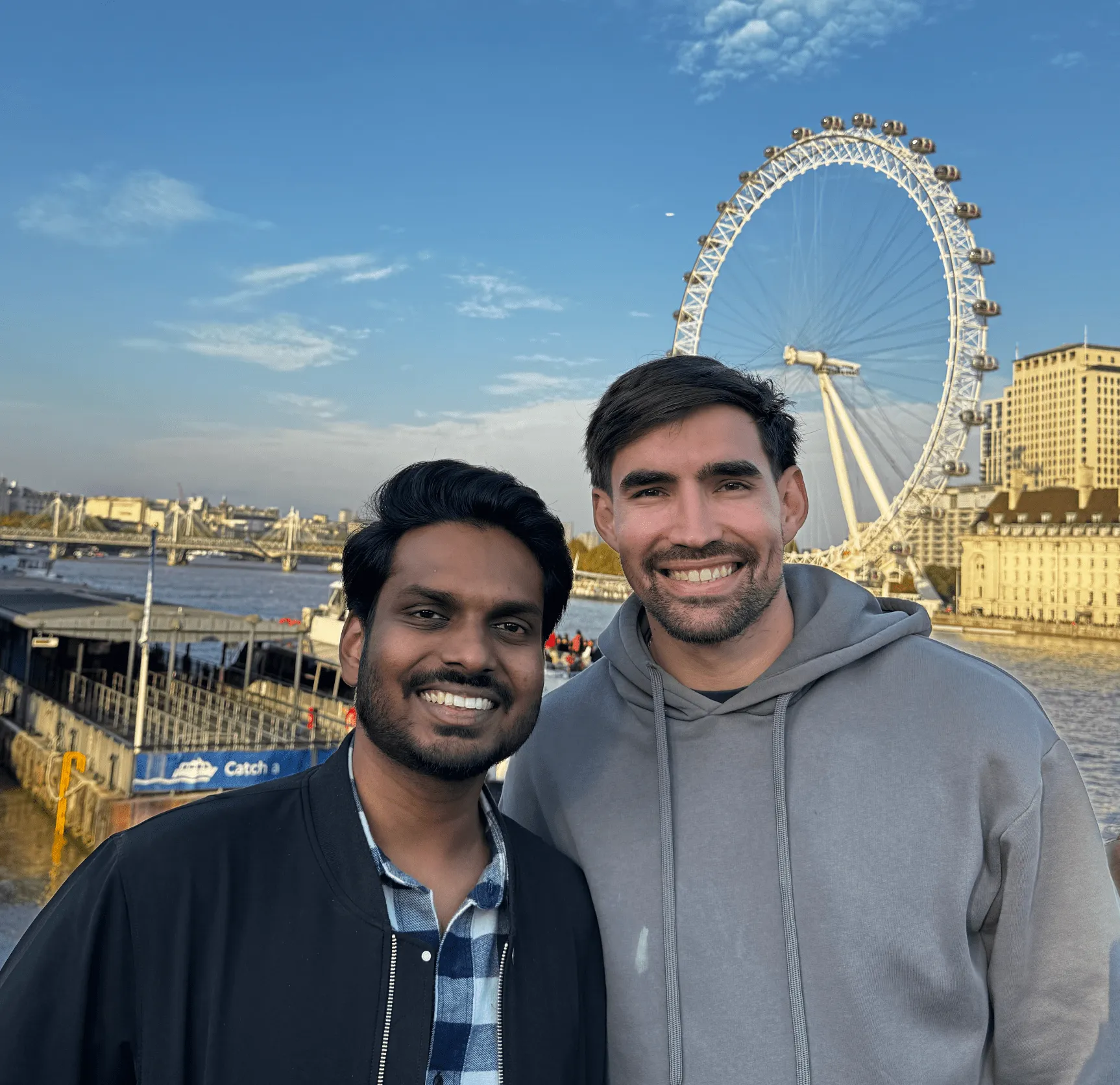 Two men smiling with the London Eye and River Thames in the background on a clear day.