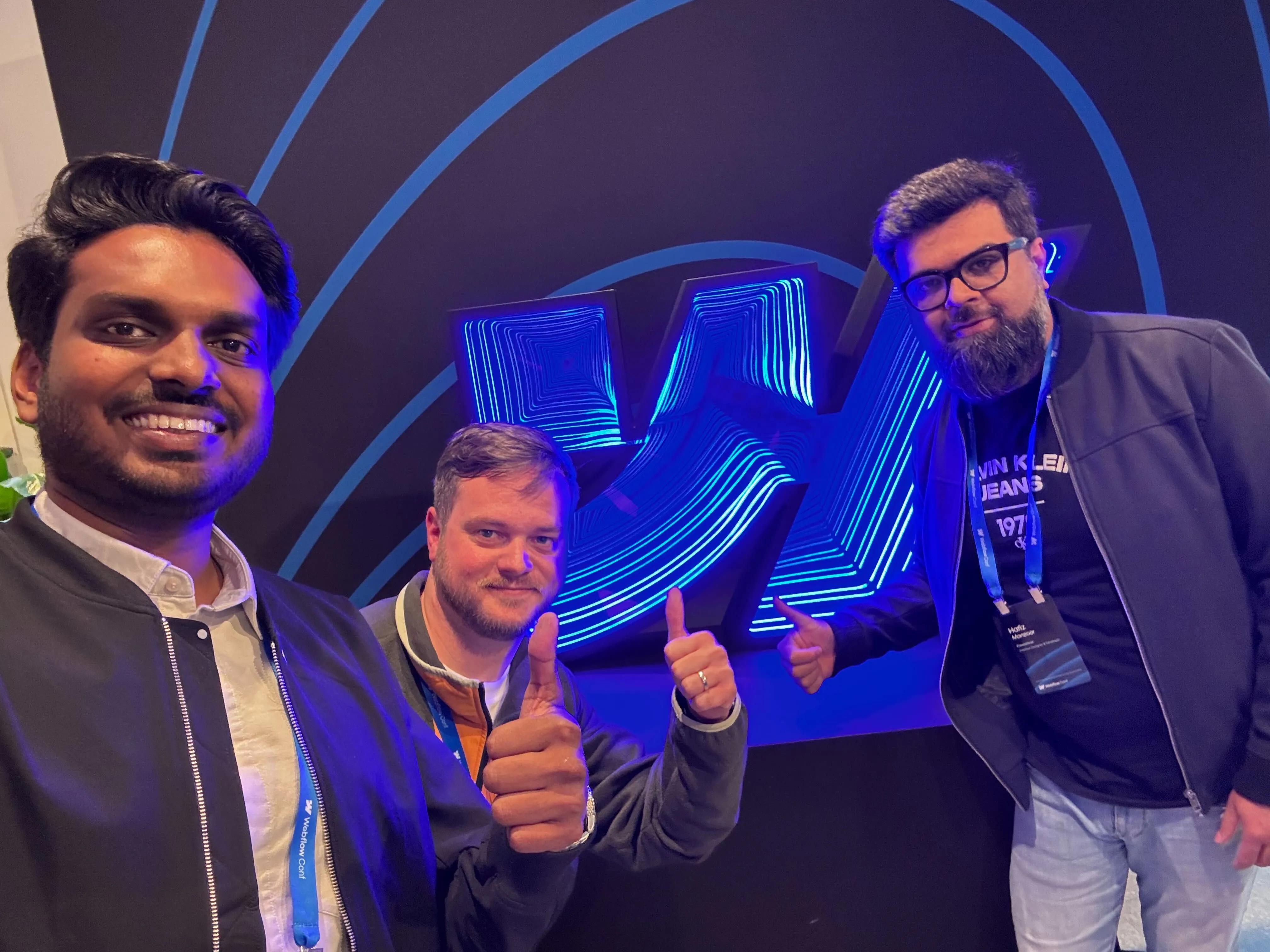 Three men smiling and giving thumbs-up in front of a blue neon abstract logo on a dark wall.
