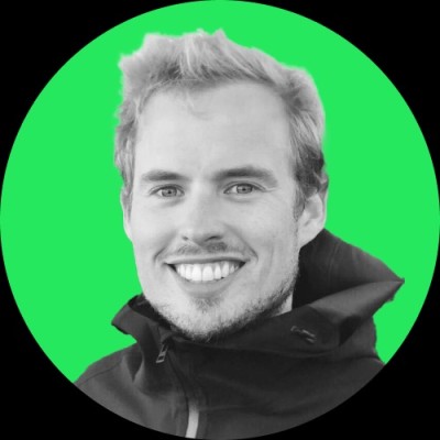 Smiling man wearing a blue suit jacket, white shirt, and light green tie standing in front of leafy green trees.