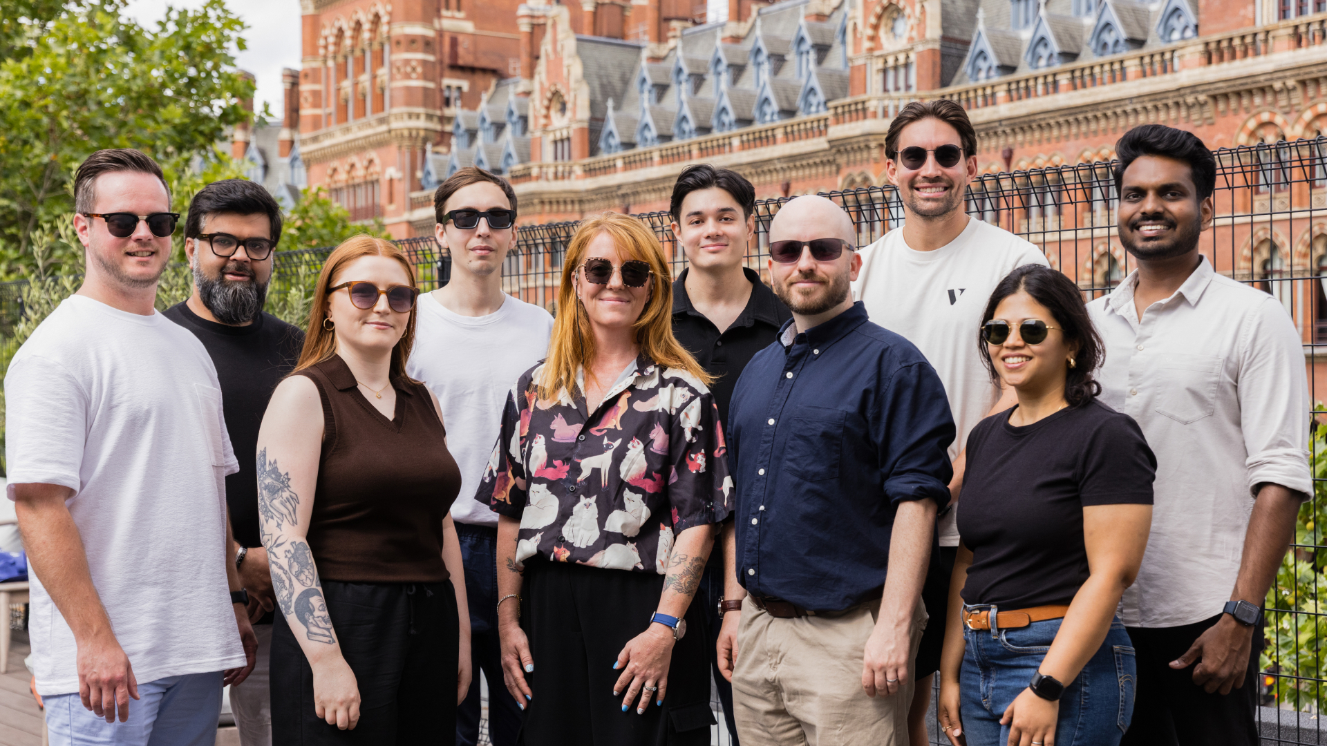 Group of nine adults standing outdoors in front of brick building, all wearing sunglasses and smiling.