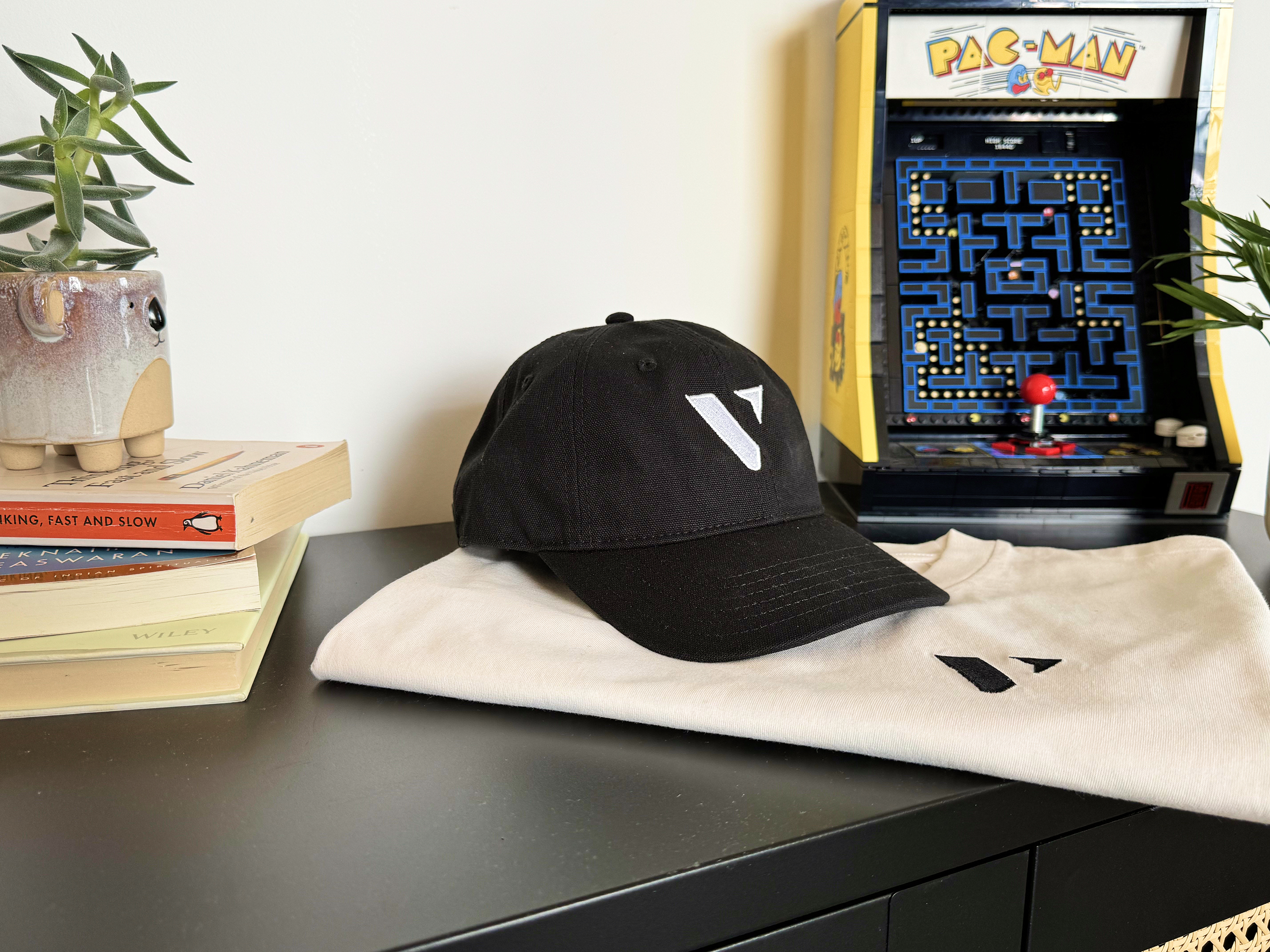 Black cap with white logo on folded white shirt beside Pac-Man arcade game and potted plant on black table.