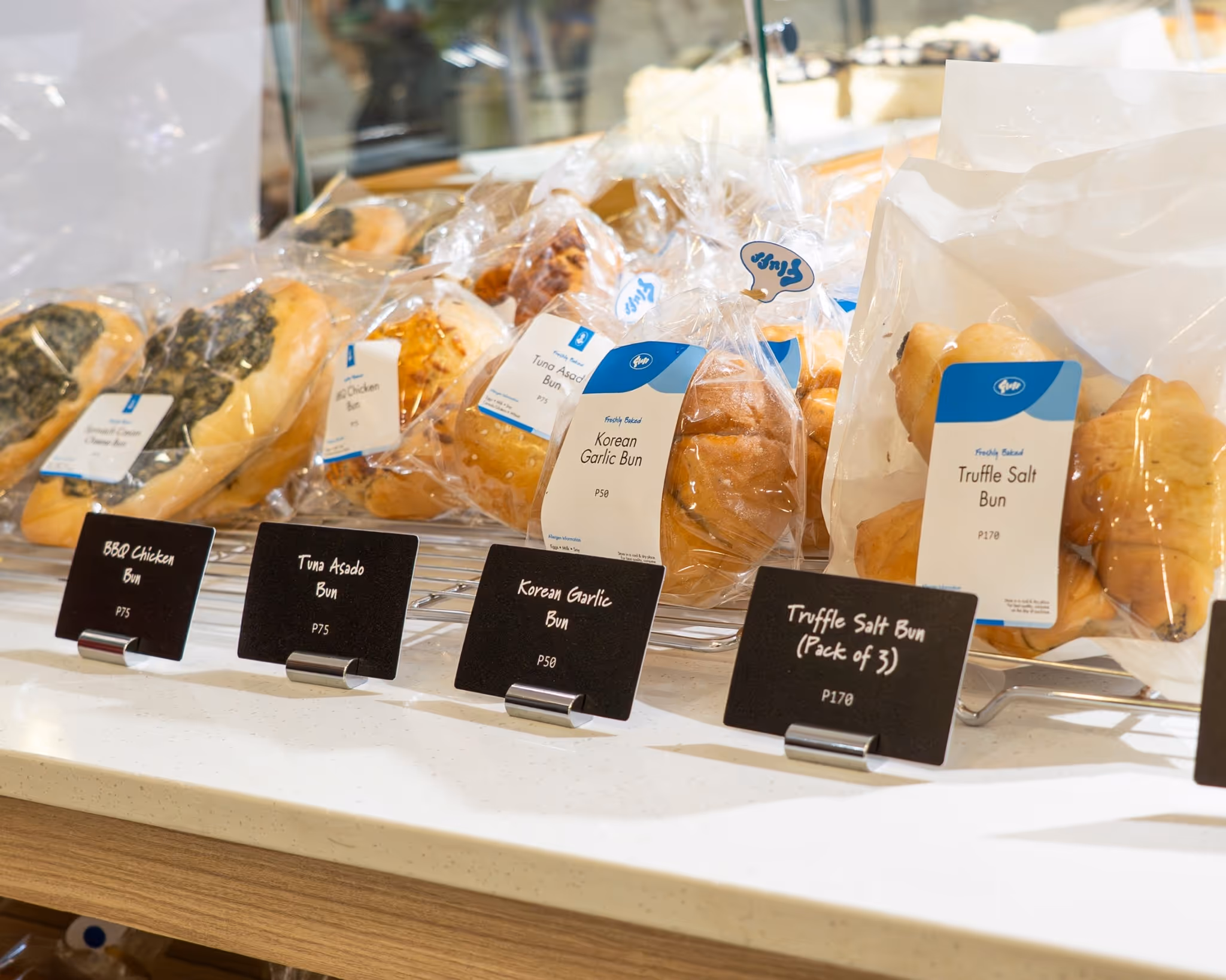 Various packaged buns including BBQ Chicken Bun, Tuna Asado Bun, Korean Garlic Bun, and Truffle Salt Bun displayed on a bakery shelf with price tags.