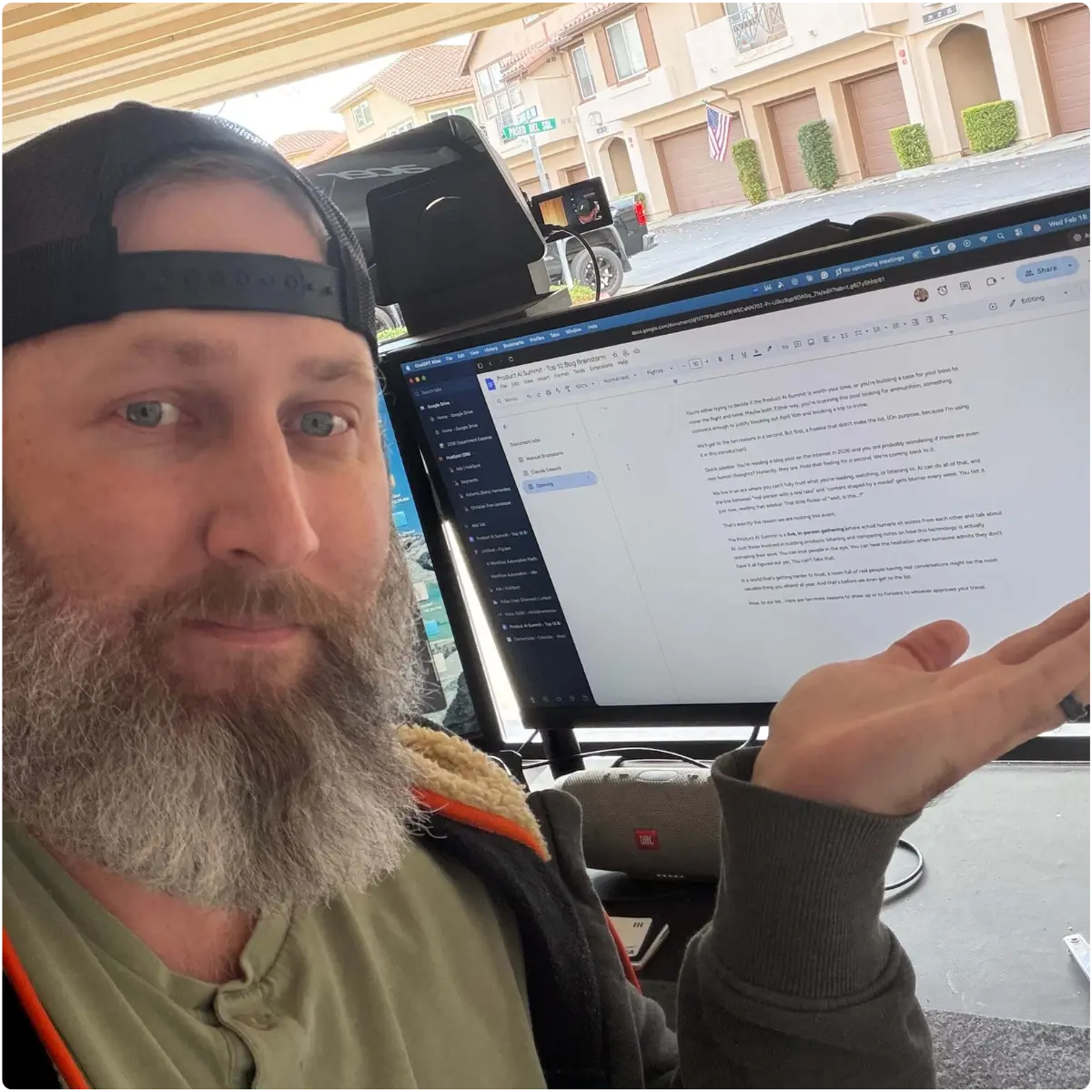 Bearded man (Chris R.) wearing a backward cap sits at a desk in a garage workspace, gesturing toward a large monitor displaying a Google Docs article draft, with a suburban neighborhood visible outside.
