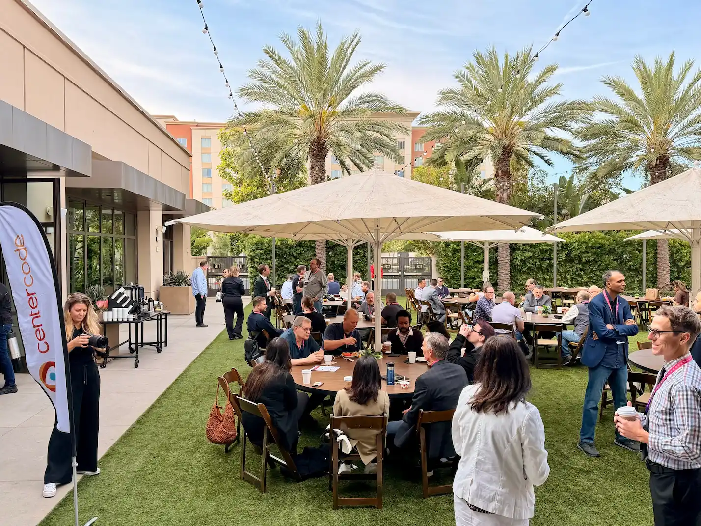 Summit attendees mingle and eat at round tables under large umbrellas on a turf patio, with a Centercode banner and coffee station to one side.