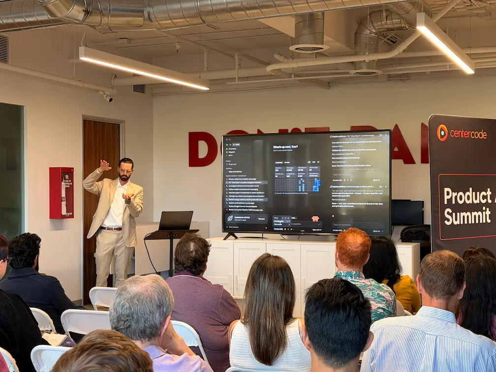 Travis Johnson in a light suit gestures to a large screen showing a Claude Code interface during a Product Summit workshop session.