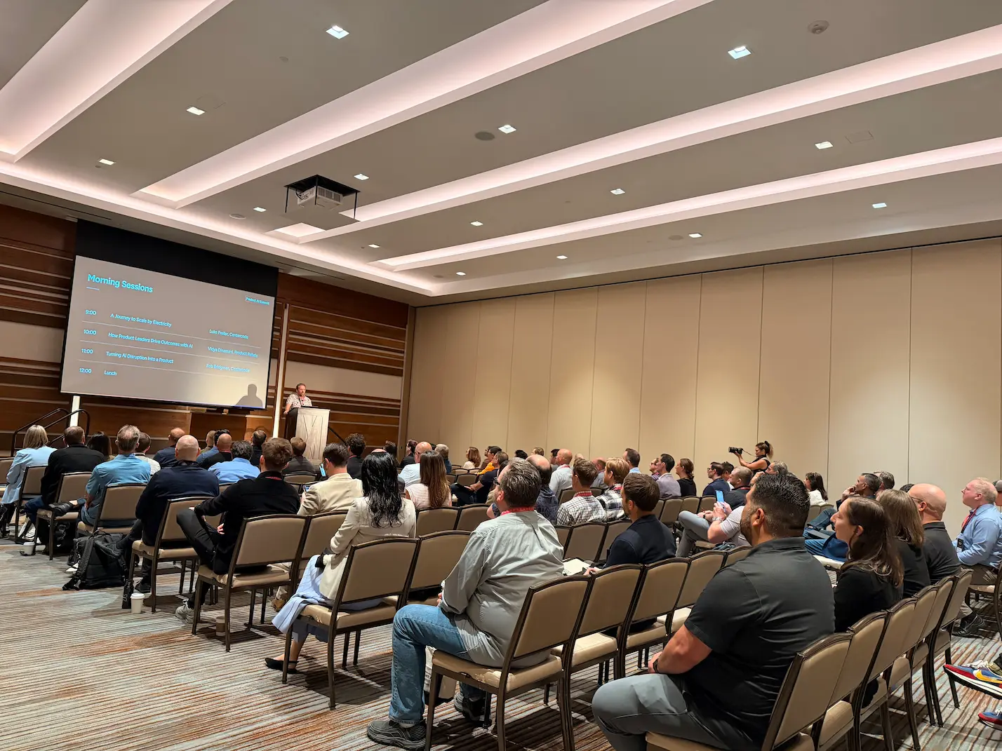 Luke Freiler addresses a seated audience from a podium in a conference ballroom, with a "Morning Sessions" agenda displayed on the large screen beside him.