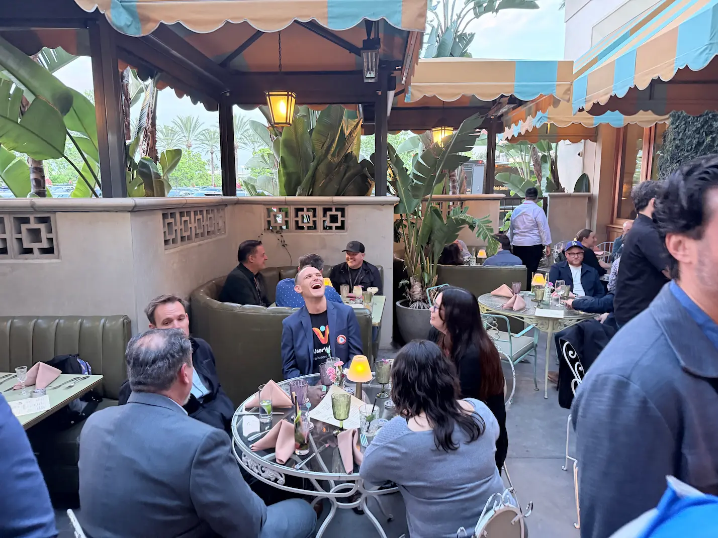  A group of Centercode attendees laughing and dining together on a lush outdoor restaurant patio with striped awnings and tropical plants.