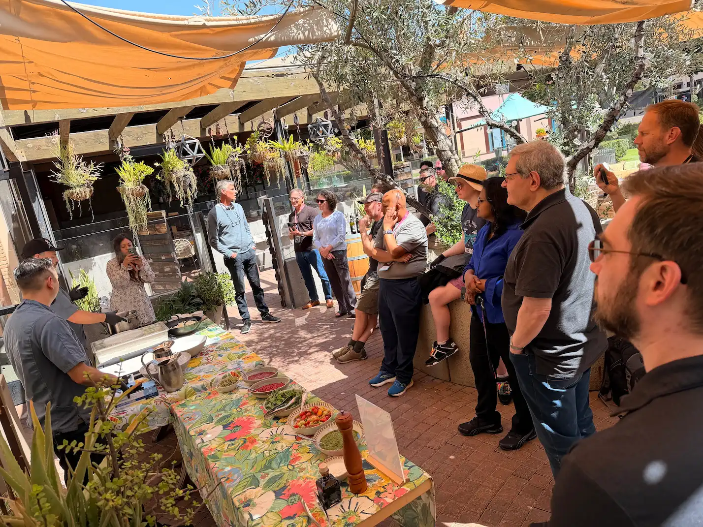 A chef demonstrates how to make fresh mozzarella to an attentive group of attendees gathered around an outdoor prep table set with fresh ingredients under shade sails.
