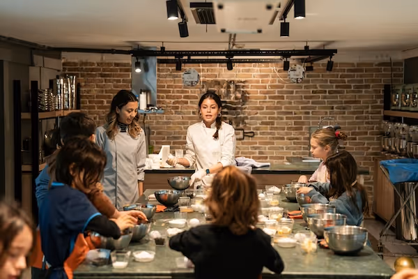 A female chef in chef whites, leading a cookery class with 5 young people.