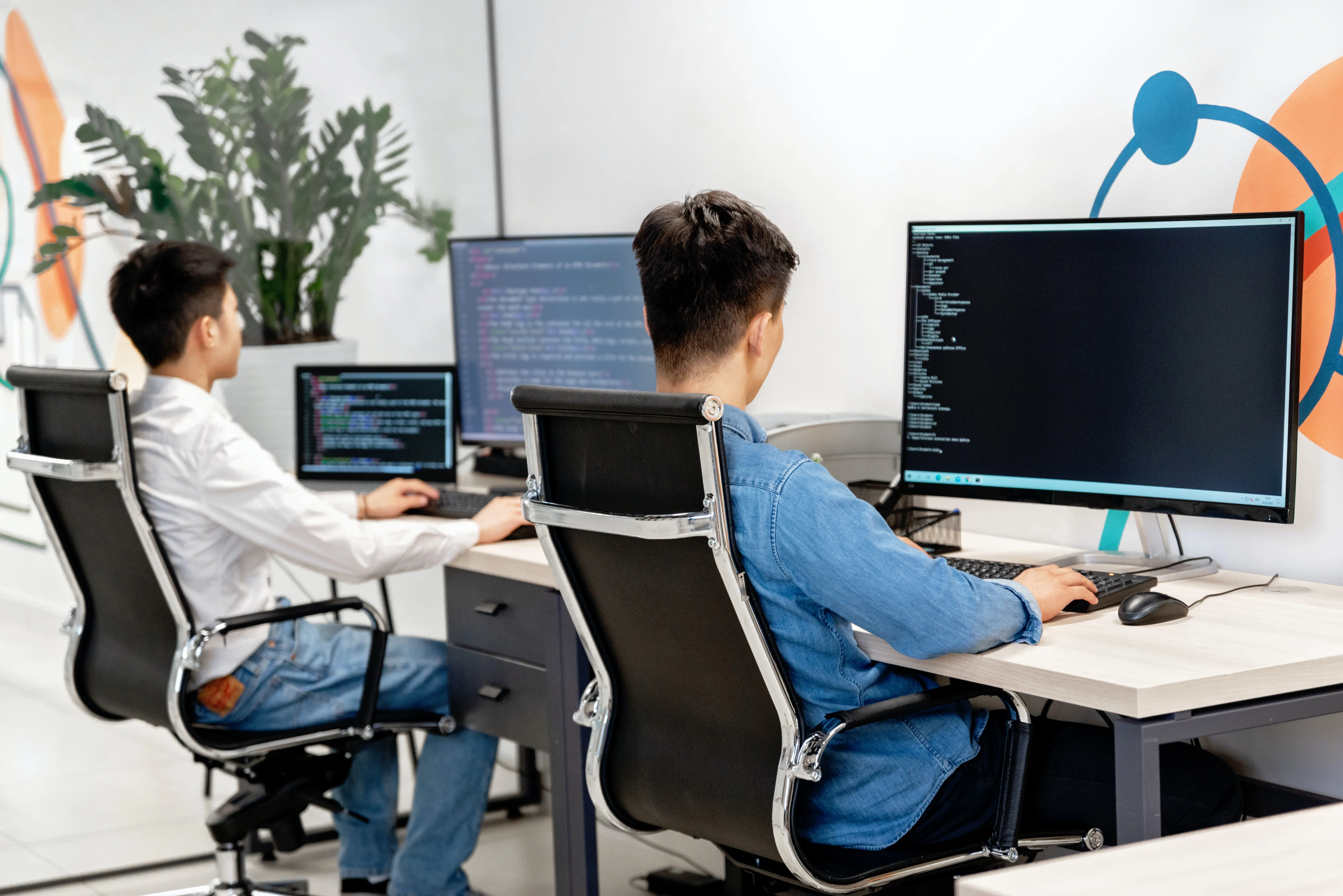 Two men sitting working at a computer desk with different IT coding on their screens