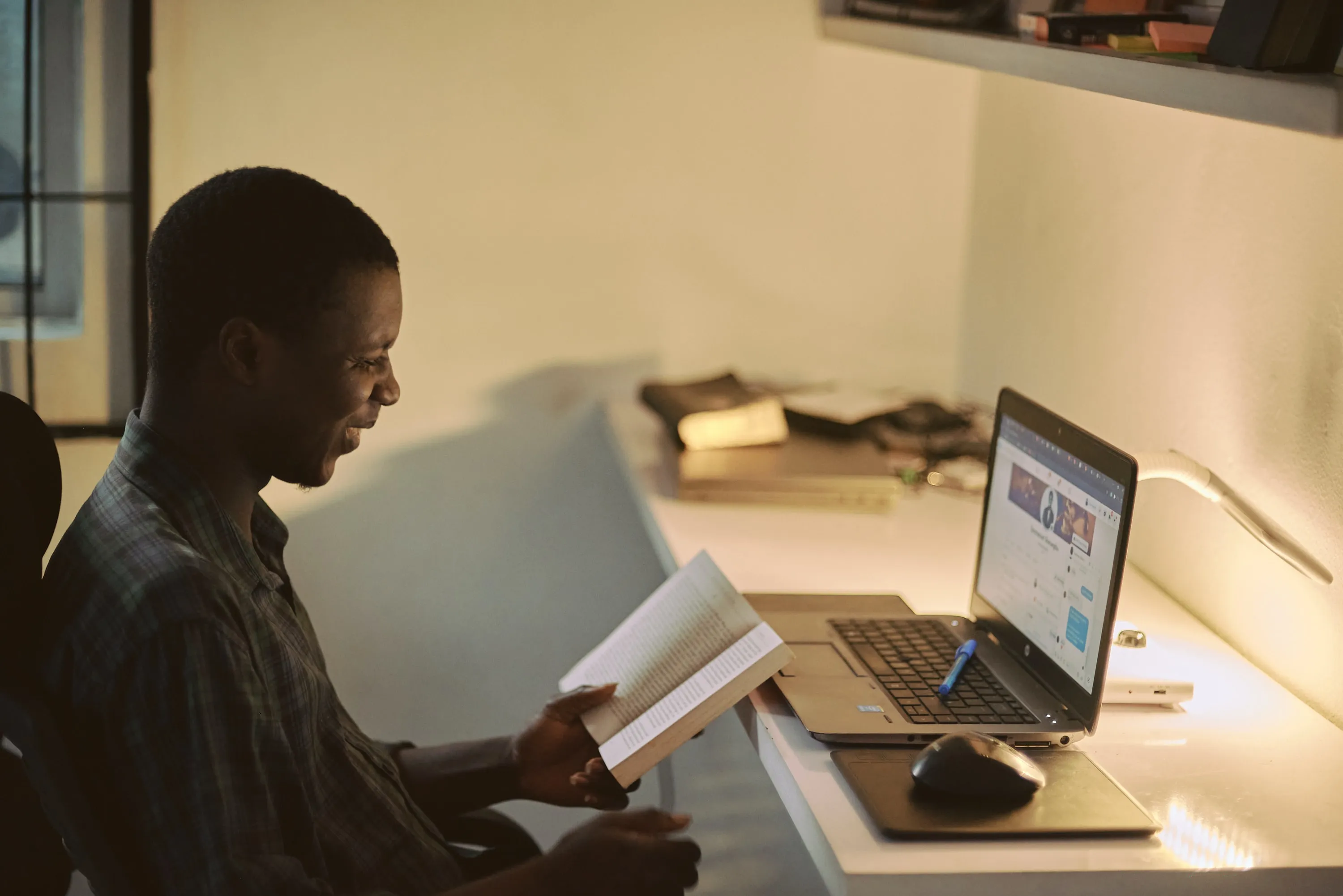 Man sitting reading a book at a desk with a open laptop