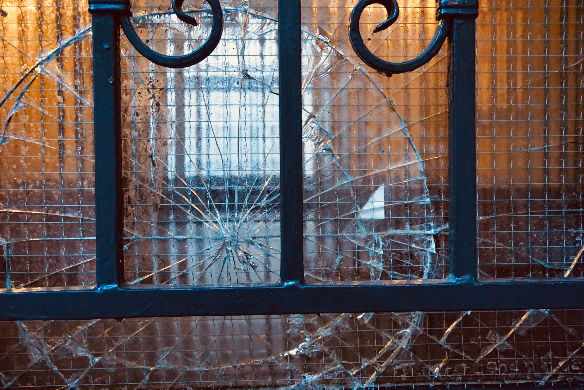 Detailní záběr drátěného skla okna s velkou kruhovou prasklinou a roztříštěnými oblastmi za kovovým rámem.