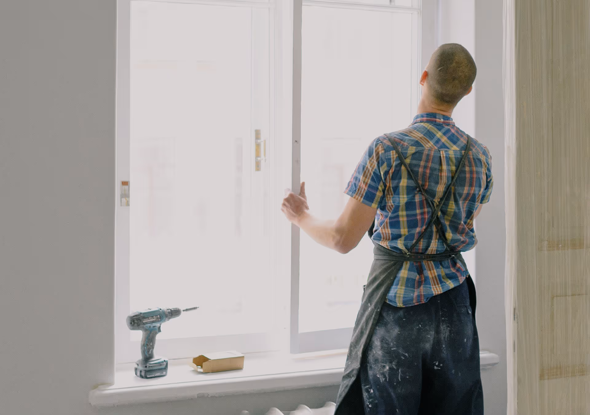 Osoba v kostkované košili a zástěře otevírající nebo instalující okno v světlé místnosti, s vrtačkou a malou krabičkou na okenním parapetu.