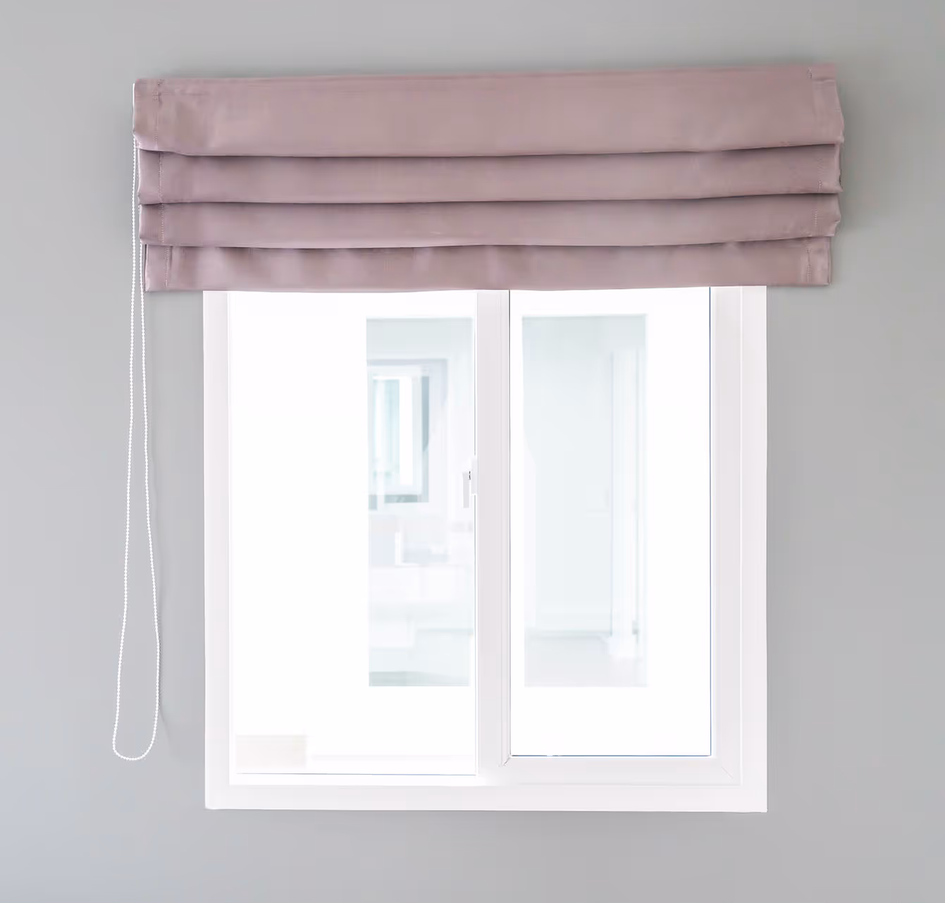 Zavřené okno s bílým rámem a složenou fialovorůžovou římskou roletou s tahací šňůrkou na šedé stěně.