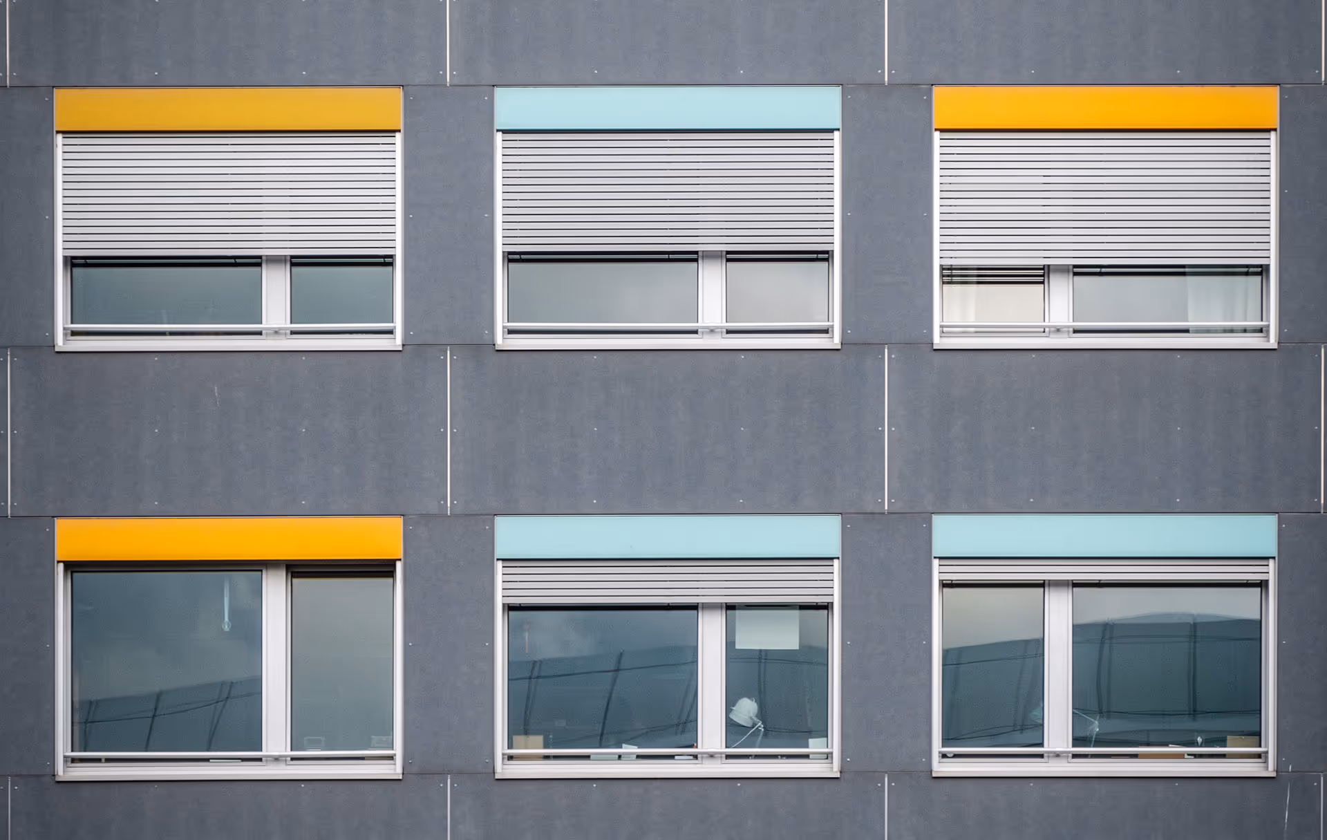 Vnější pohled na šest moderních obdélníkových oken s šedými roletami na tmavě šedé stěně budovy; tři okna mají v horní části oranžové panely a tři světle modré panely.