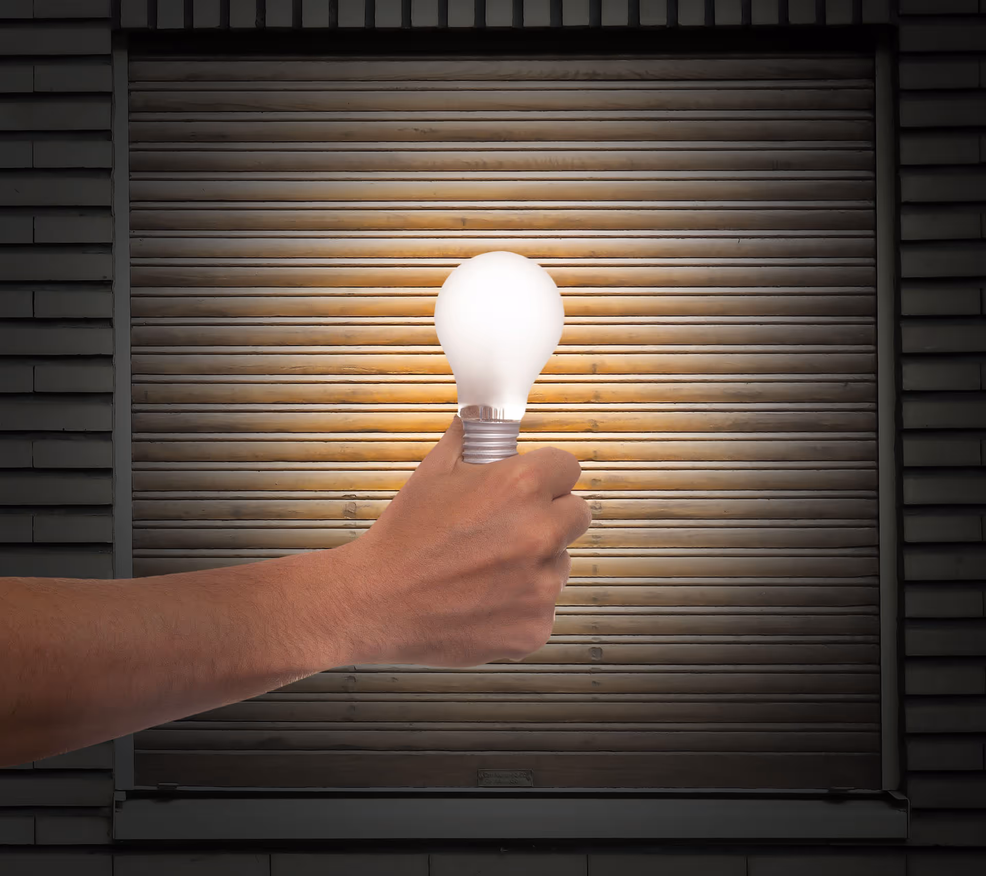 Ruka držící svítící žárovku před zavřenou kovovou roletou.