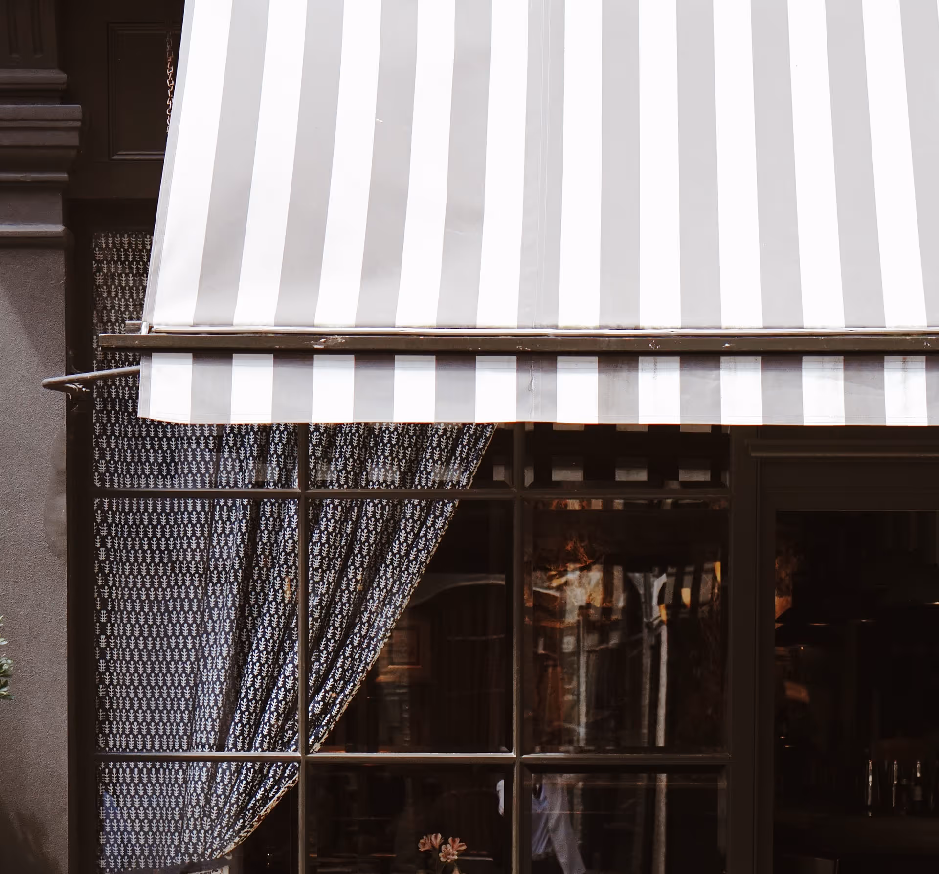 Šedobílá pruhovaná markýza nad oknem se vzorovanými záclonami a odrazy ve skle.