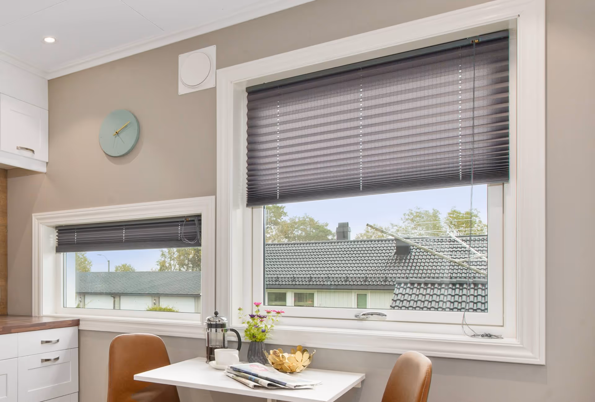 Moderní kout kuchyně se dvěma hnědými židlemi a bílým stolem, na kterém je french press, šálek, časopis, květiny a zlatá dekorativní mísa, pod dvěma okny s částečně zataženými šedými žaluziemi.