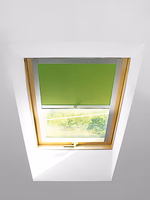 Střešní okno s částečně spuštěnou zelenou roletou a dřevěným rámem, nabízející výhled na stromy venku.
